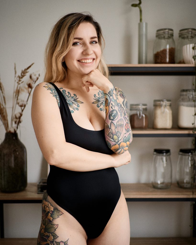 Kuvassa Viivi Kohtanen. Tatuoitu nainen hymyilee kameralle keittiön hyllyn edessä. Objektiivin Sony FE 35mm f/1.8 testikuva. Valokuva: Anssi Lepikko.